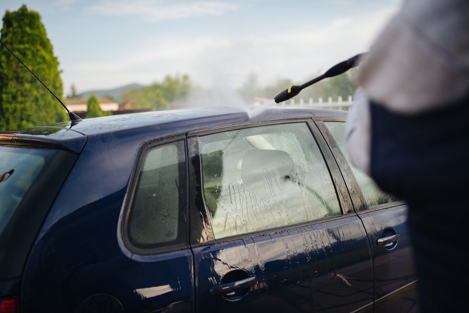 A pressure washer rinsing suds from the roof and side glass of a dark blue hatchback outdoors