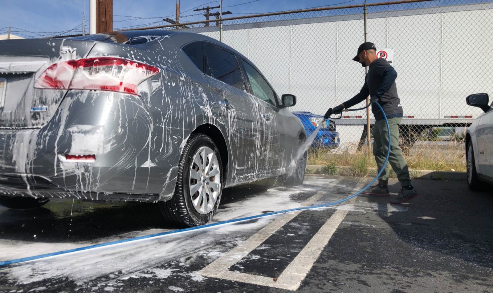 A detailer rinsing foam from a soapy grey sedan on location, hose running across the ground beside the car